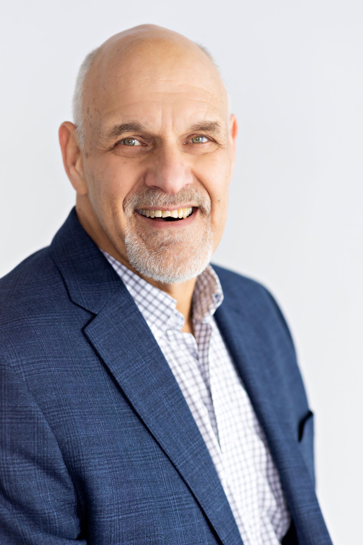 Smiling older man with a beard, wearing a blue blazer and checkered shirt, against a light background.