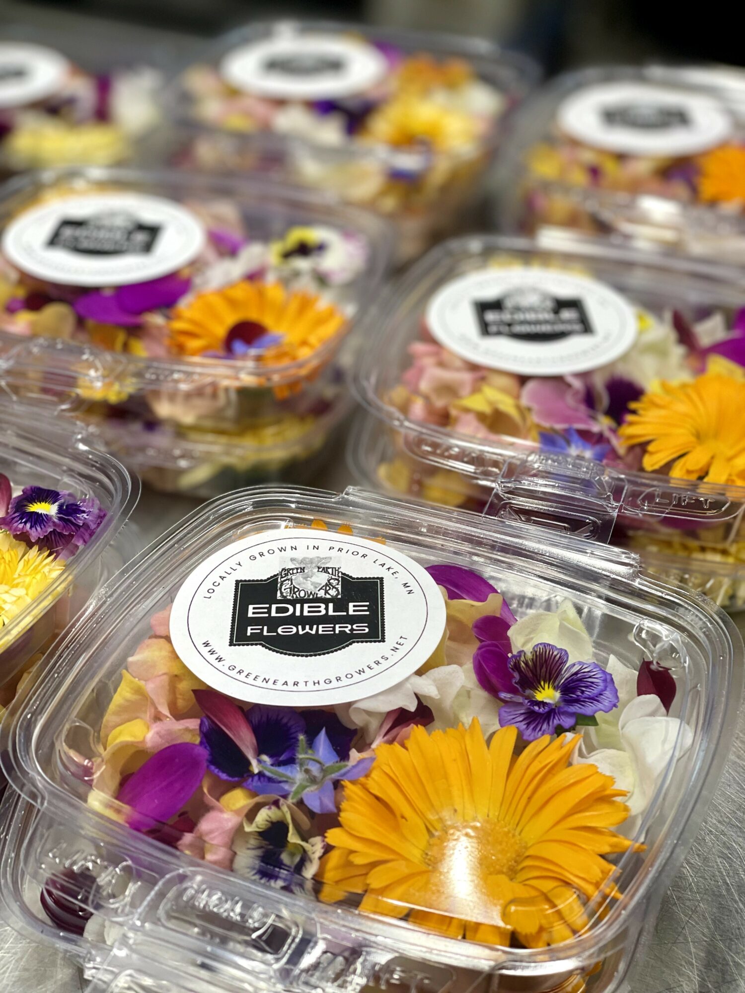 Clear plastic containers filled with colorful edible flowers including yellow, purple, and white petals, labeled 'Edible Flowers'.