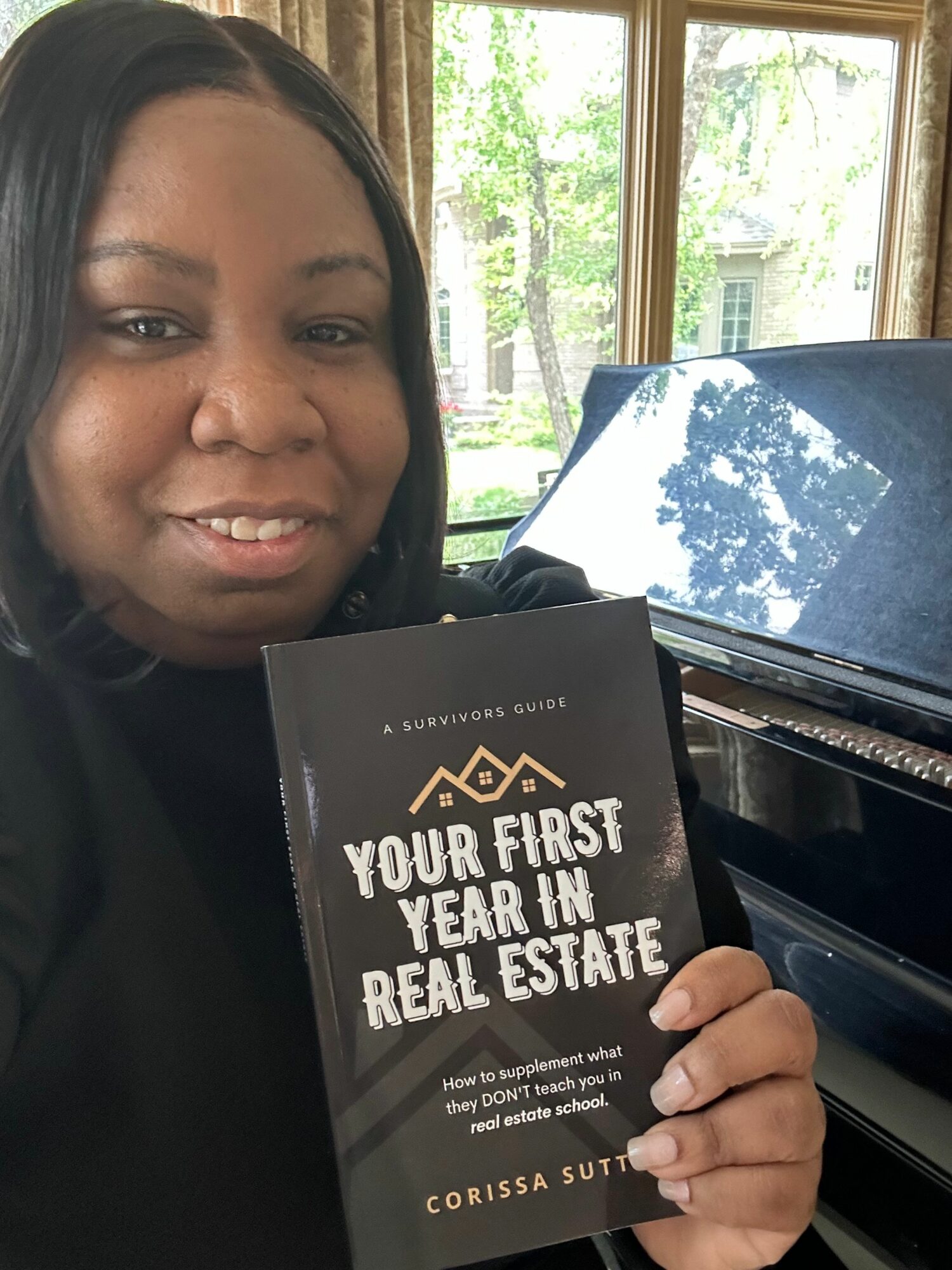 Woman holding a book titled 'Your First Year in Real Estate' indoors near a window with a piano in the background.