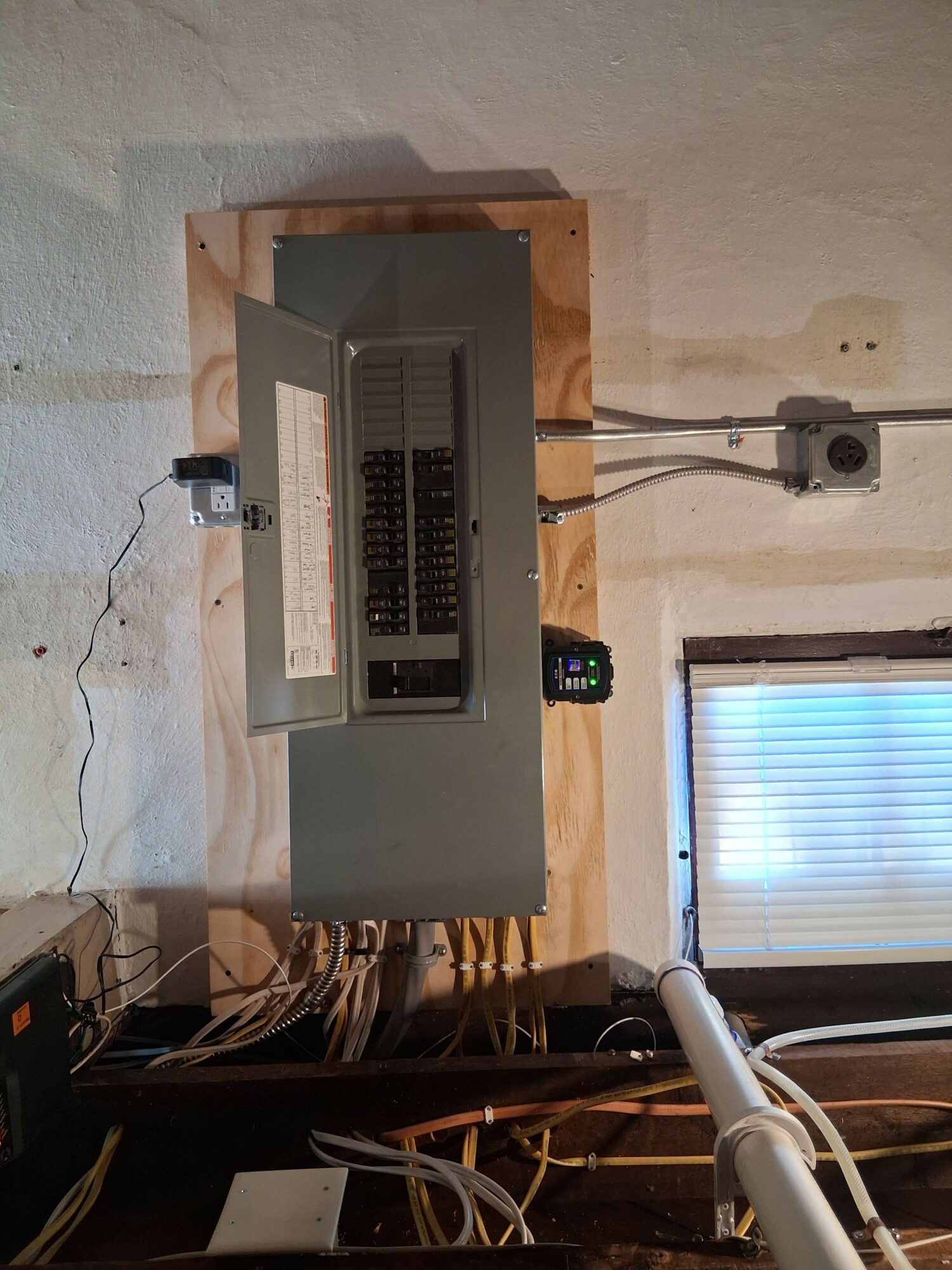 Electrical panel with wires and a small digital device mounted on the wall near a window with blinds.