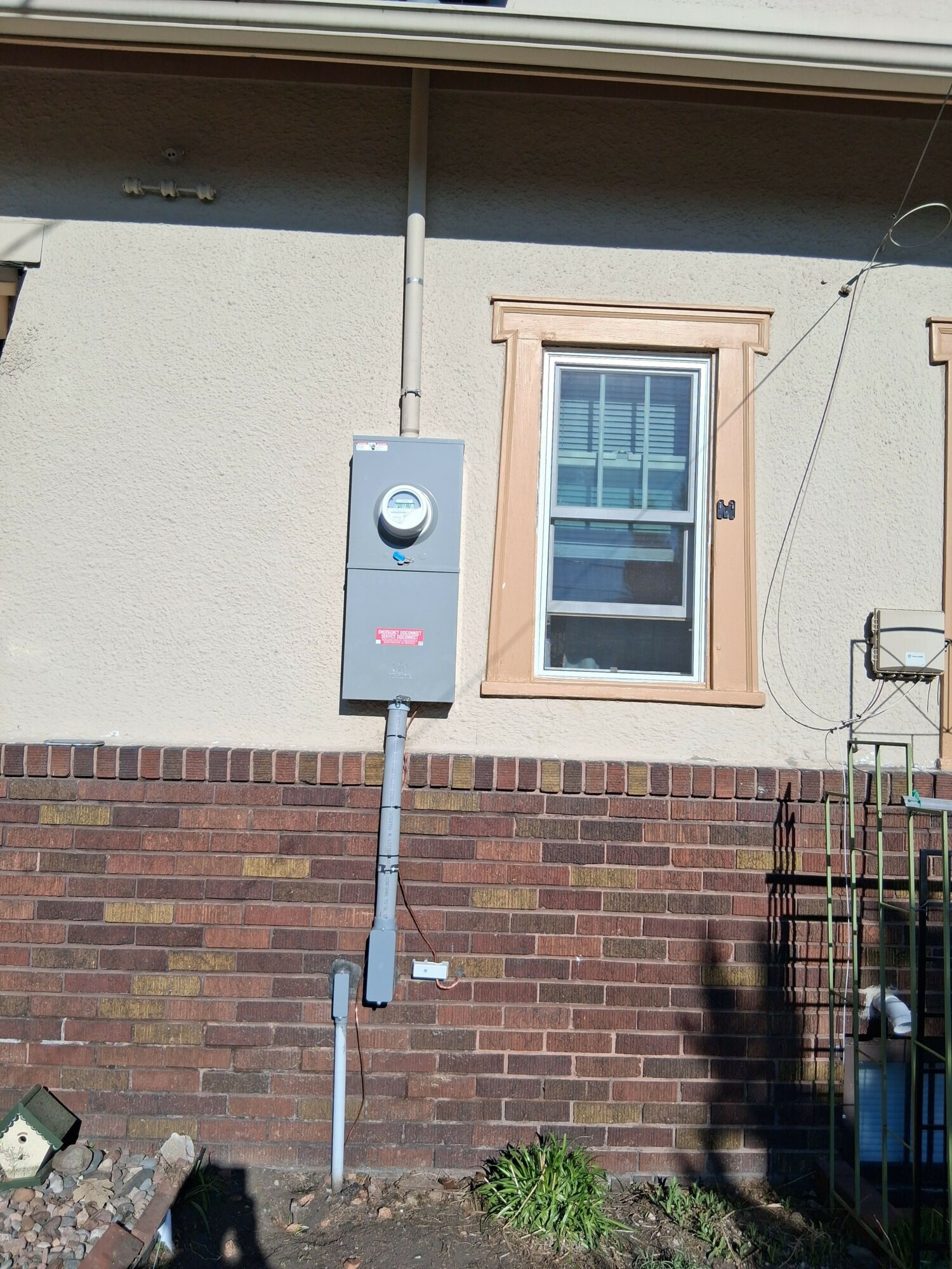 Exterior wall with window and electrical box, brick and stucco surface, small plant at base, part of a fence on right.