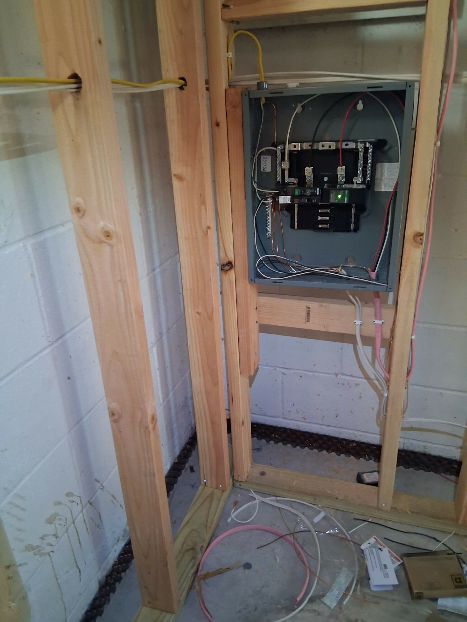 Open electrical panel with circuit breakers and wires inside a wooden frame, with wires on the floor nearby.
