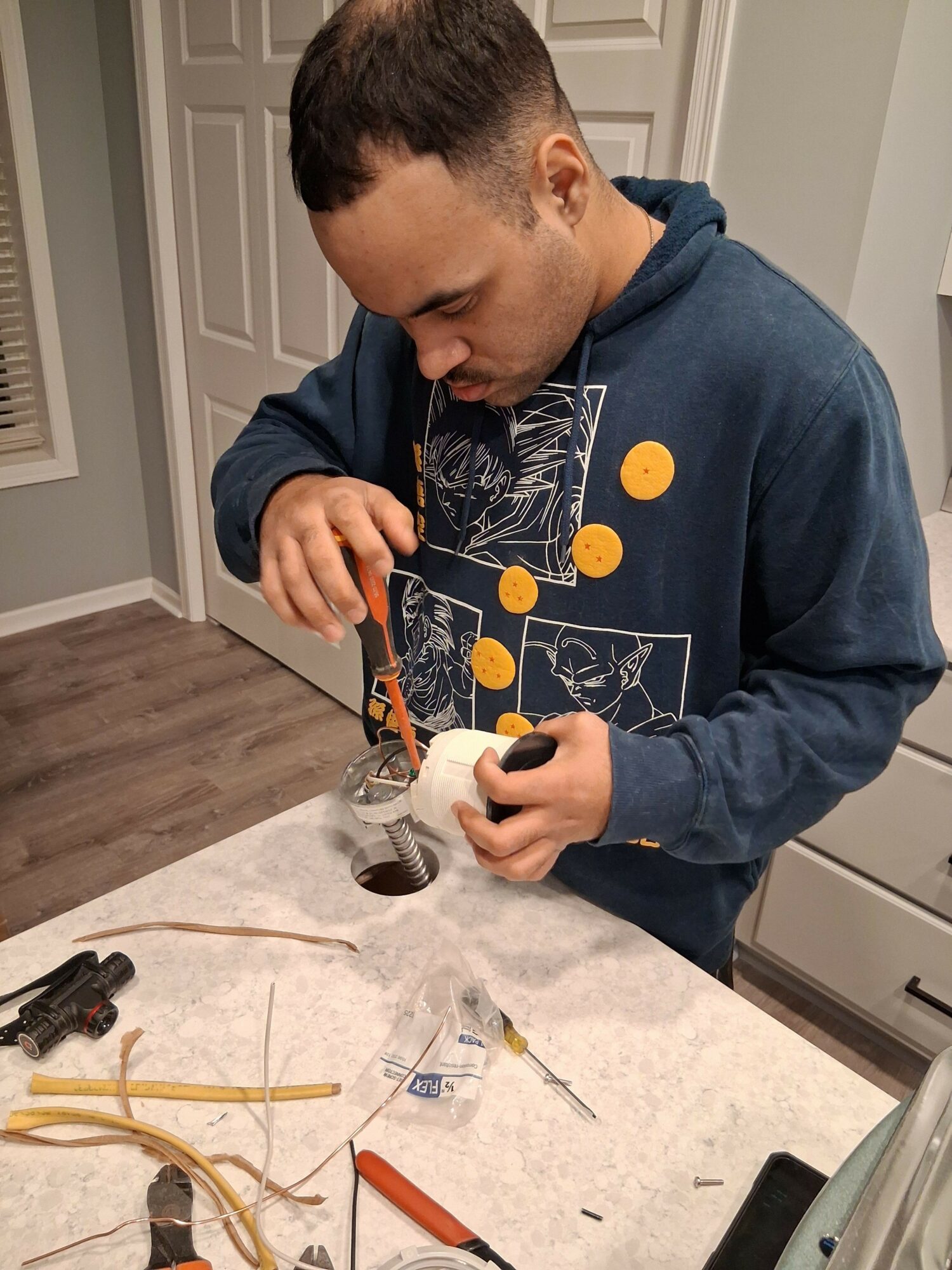 Young man in hoodie working with a small device on a kitchen counter, surrounded by tools and wires.