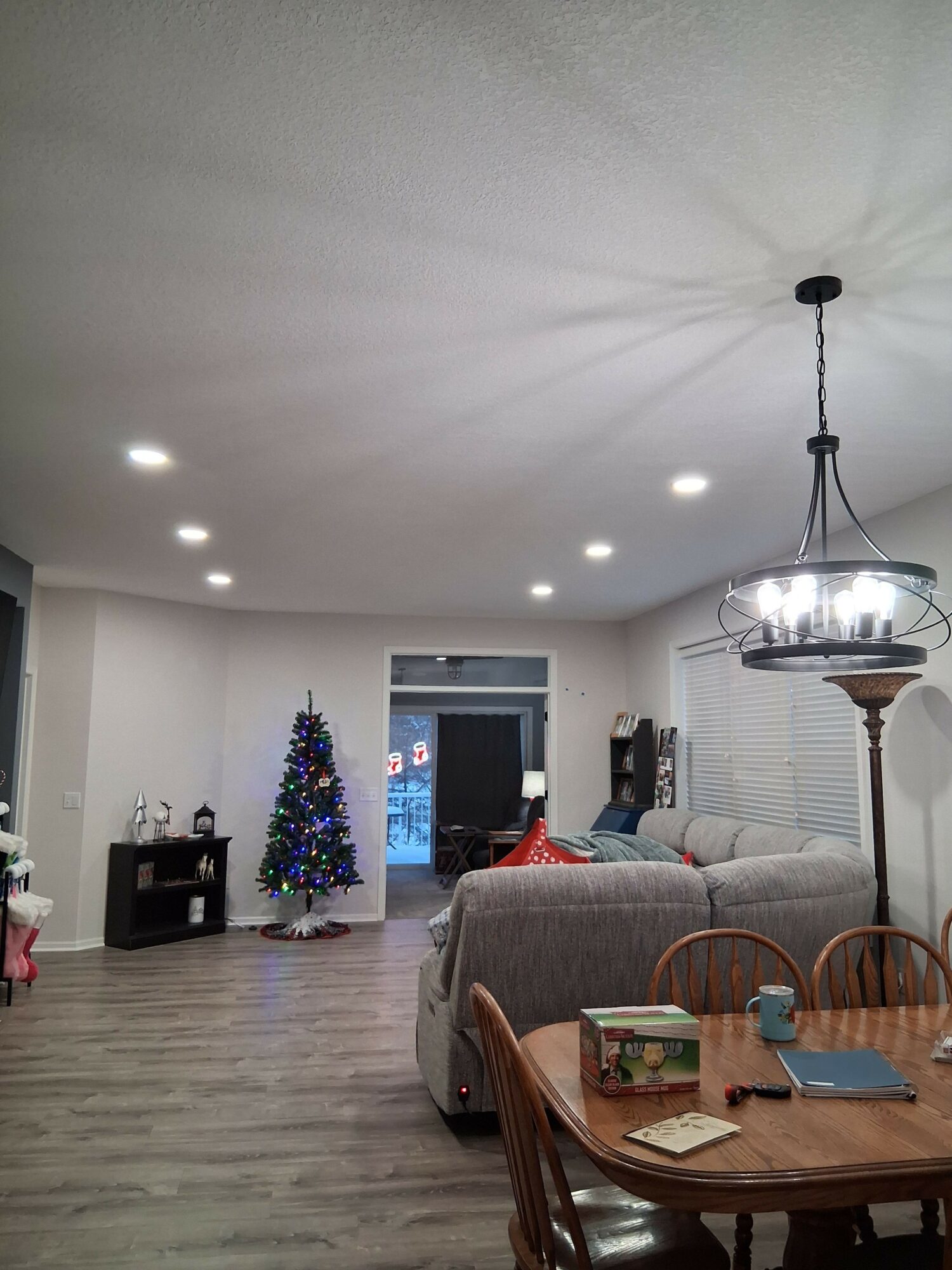 Living room with Christmas tree, sofa, dining table, and chandelier, illuminated by ceiling lights and a lamp.