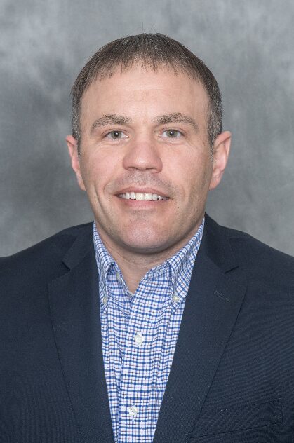 Man with short brown hair, wearing a dark blazer and blue checkered shirt, smiling against a gray background.