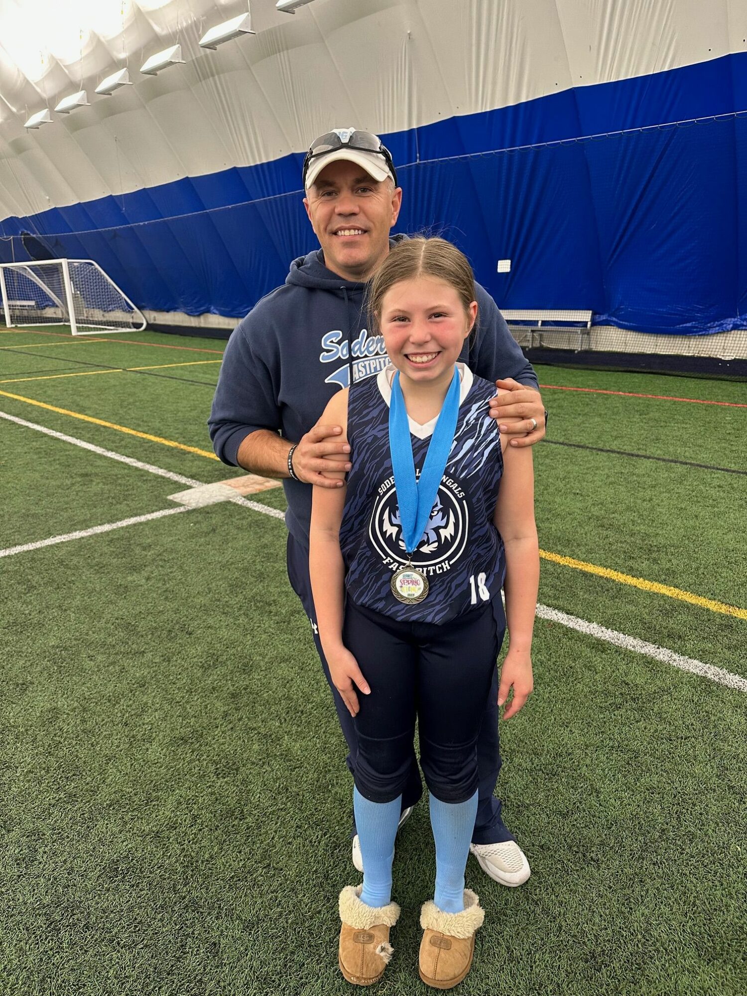 Smiling girl with a medal around her neck standing on an indoor sports field, with a man behind her.