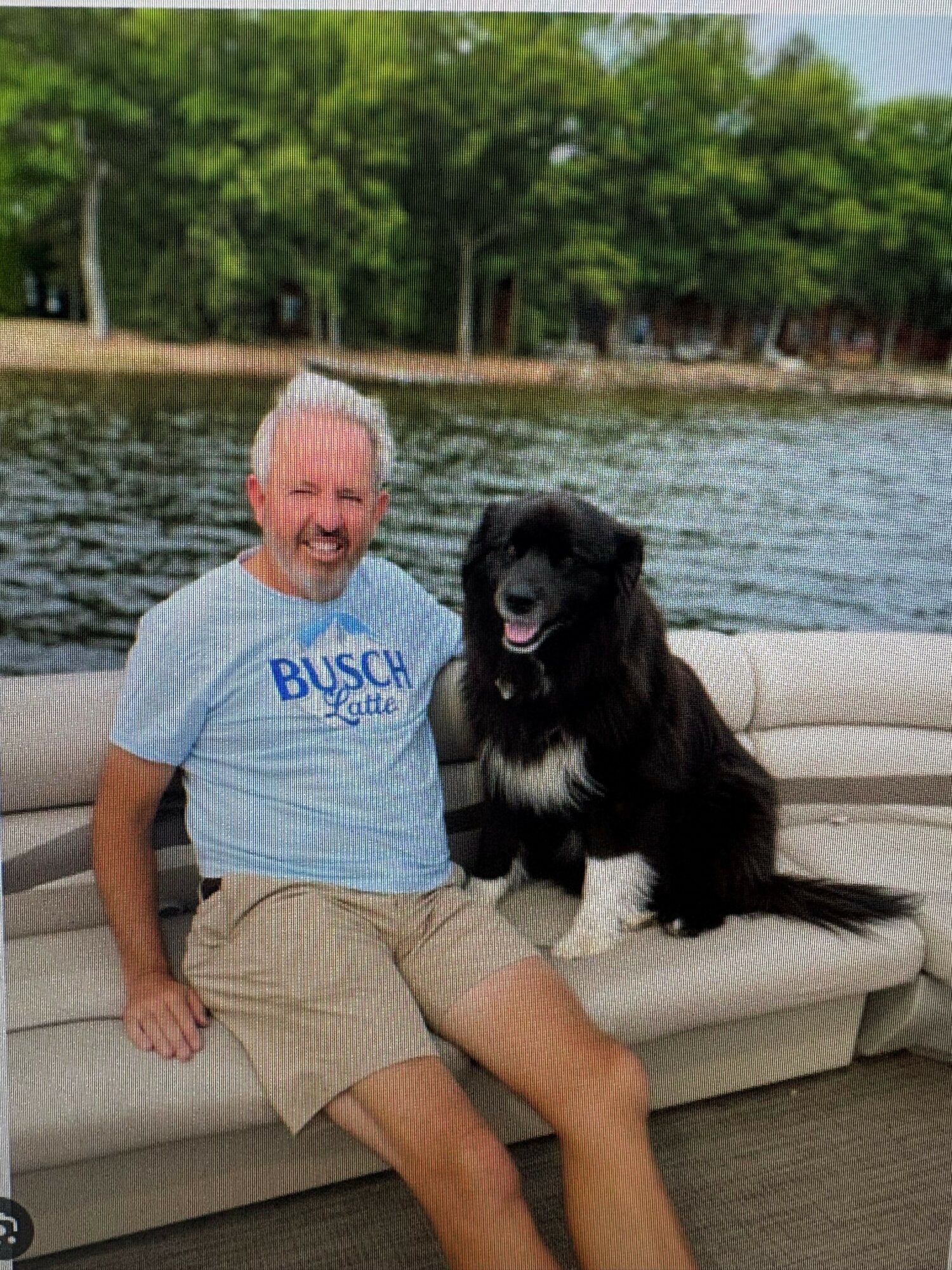 Older man with white hair and a beard sitting on a boat with a black dog, water and trees in background.