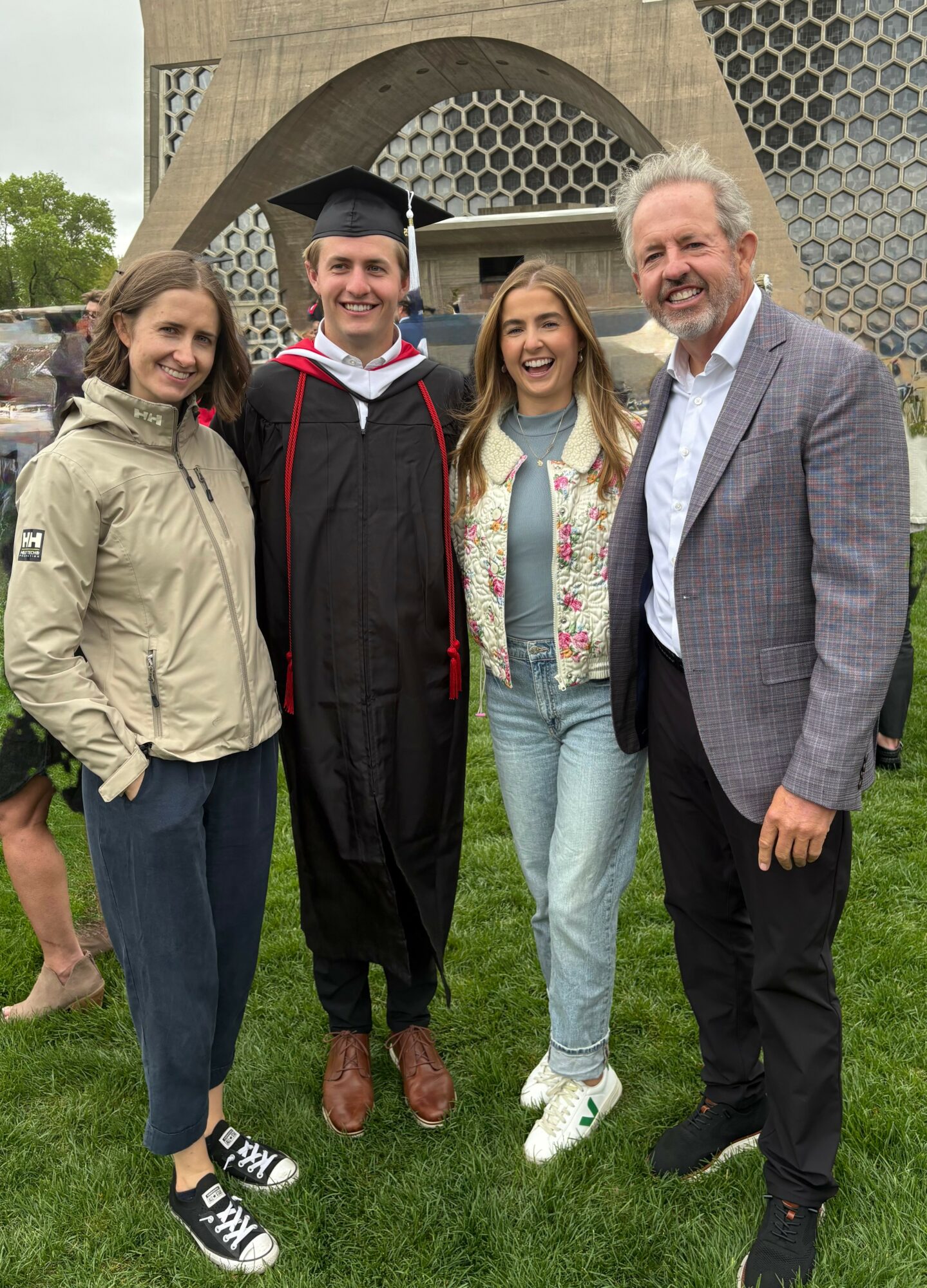 Four people standing outdoors, one in graduation gown and cap, smiling, with a modern building in background.