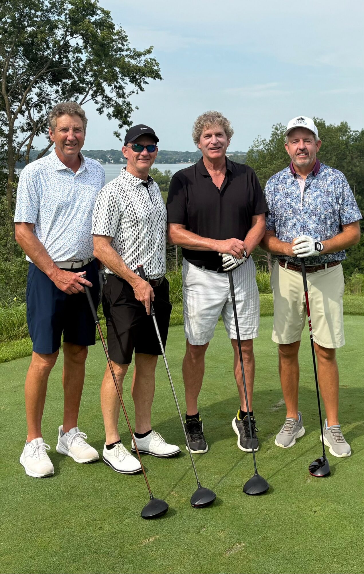 Four men standing on a golf course holding golf clubs, smiling, with trees and sky in background.