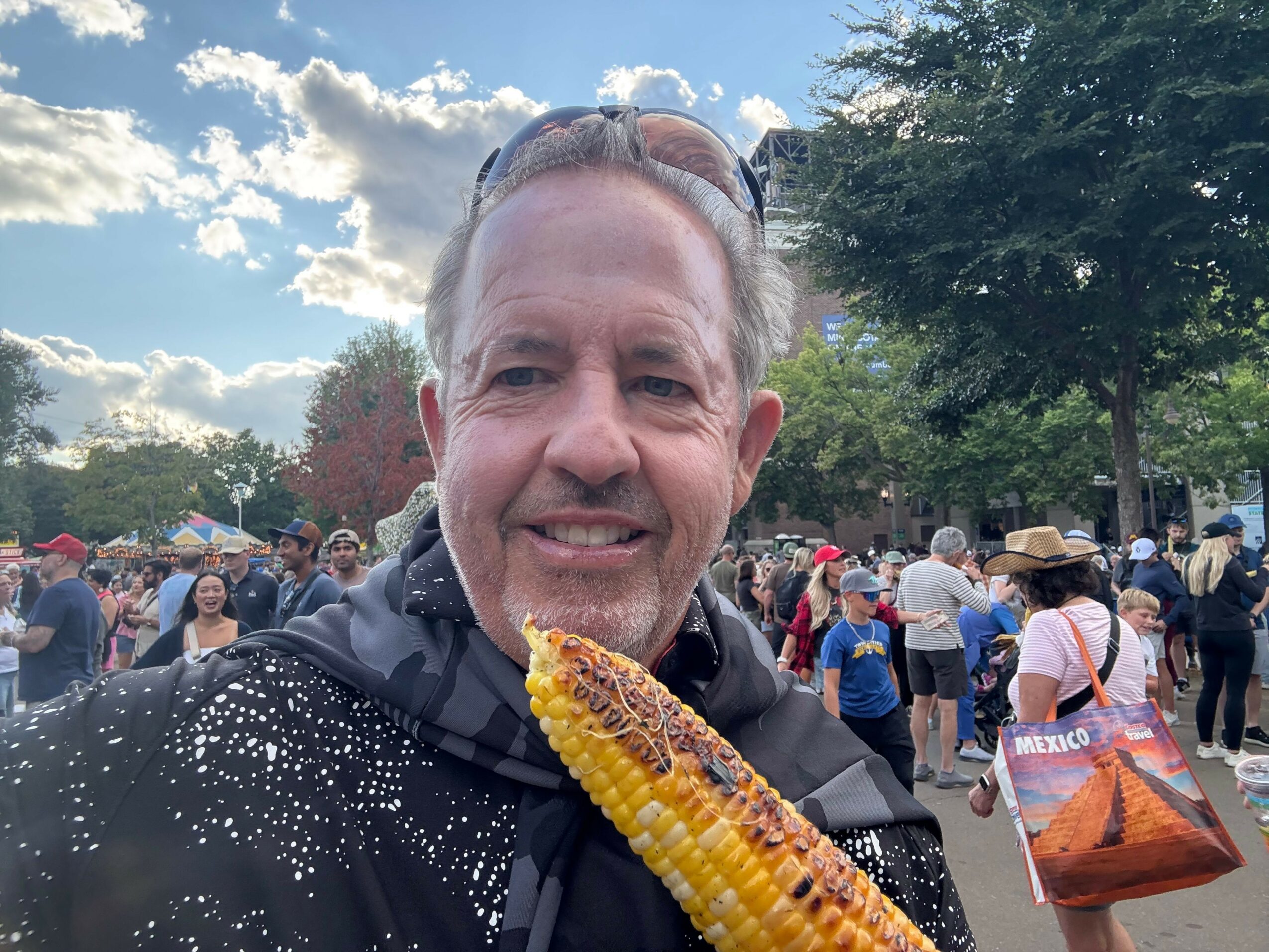 Man smiling outdoors at a crowded event with trees and clouds in background.