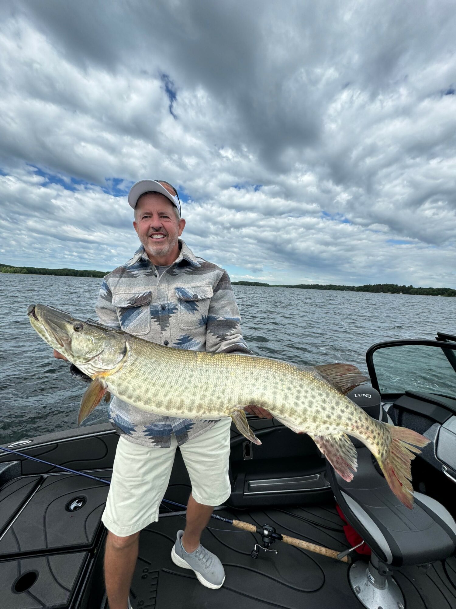 Man holding a large fish on a boat with a cloudy sky and water in the background.