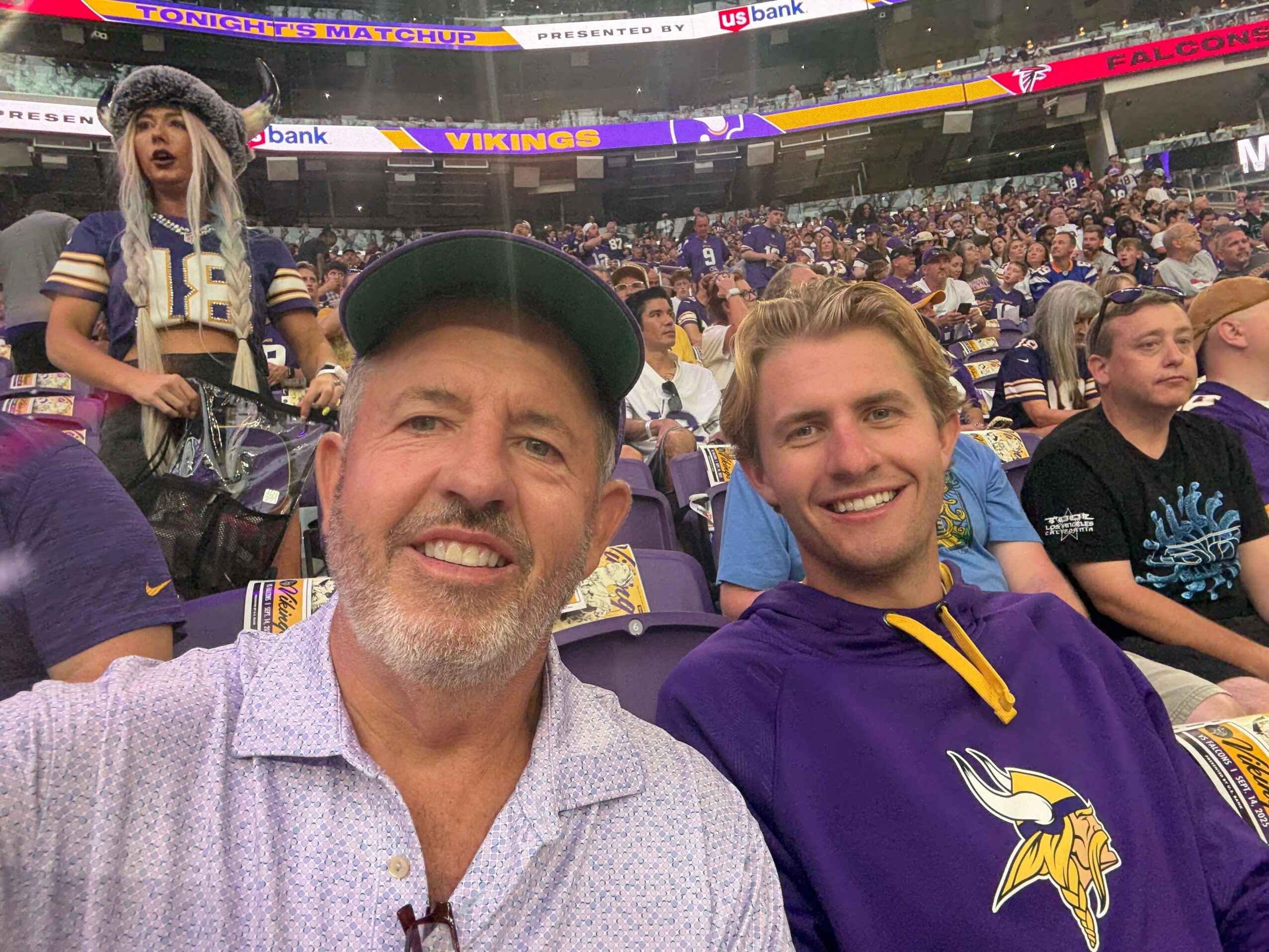 Two men smiling at a sports event, surrounded by seated spectators and a large stadium background.