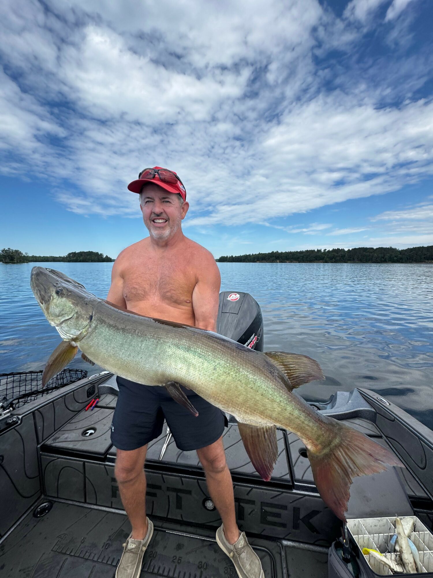 Man in red cap and black shorts holding large fish on boat, water and sky in background.