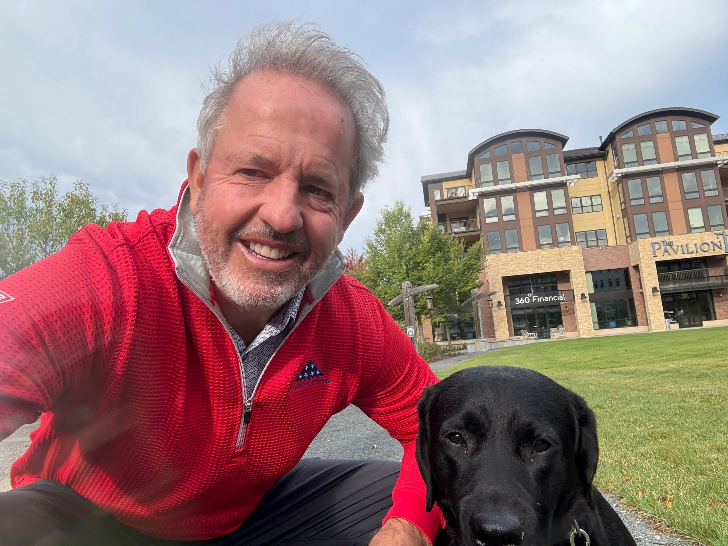 Smiling man with gray hair and beard wearing a red jacket outdoors with a black dog.