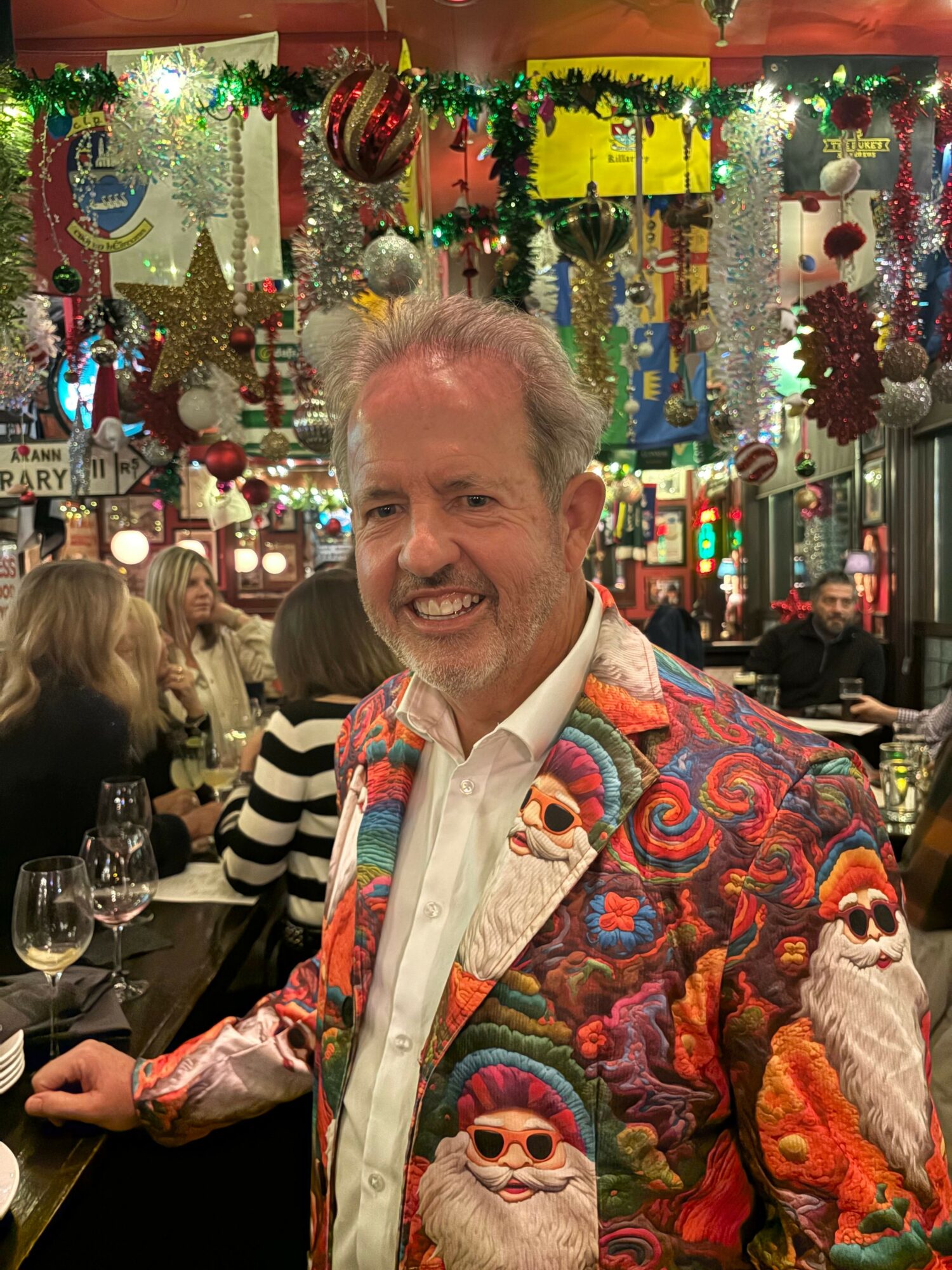 Smiling man in colorful jacket at festive indoor celebration with decorations and people in background.