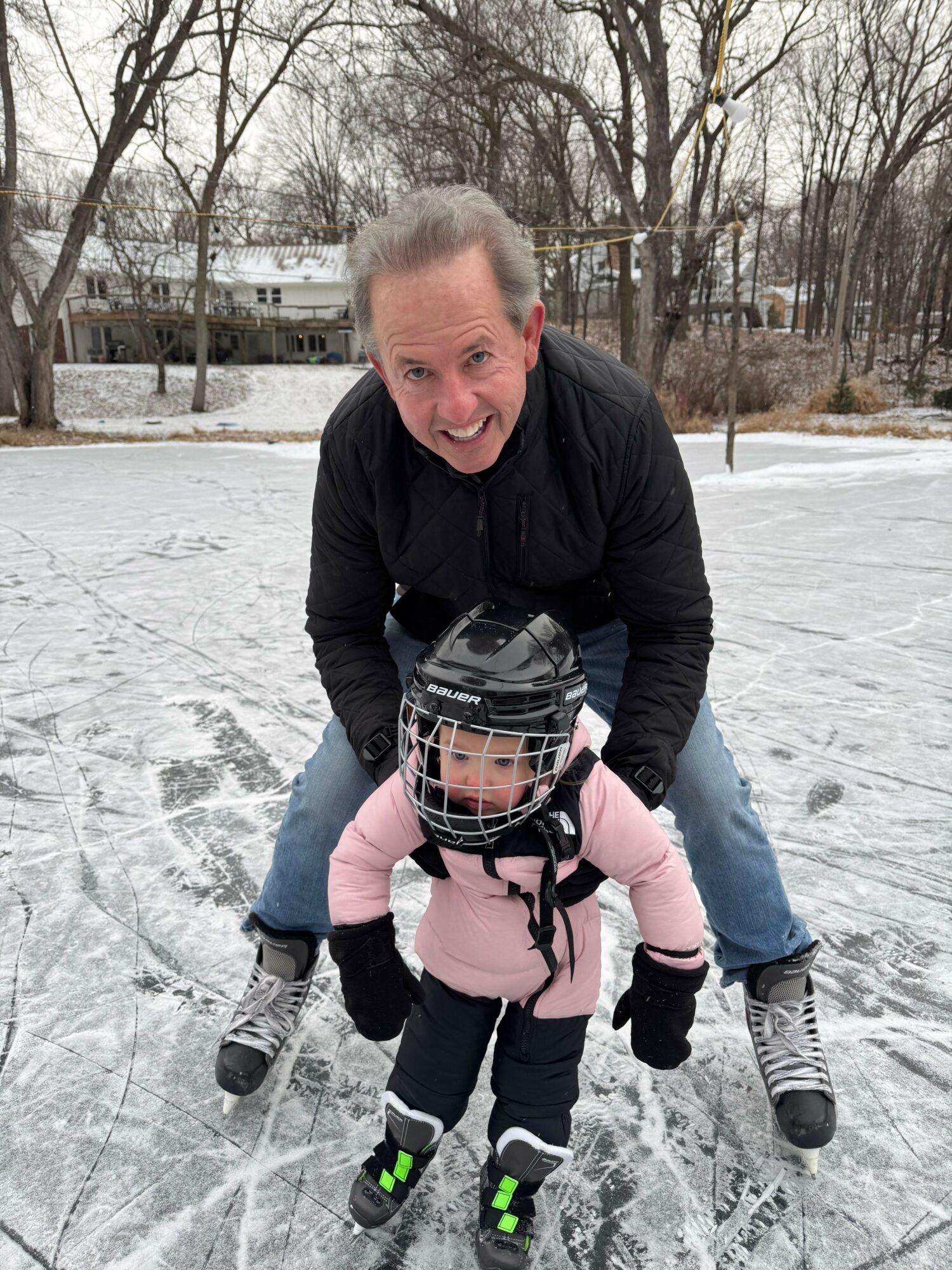 Adult and child on ice rink, adult bending over, child wearing helmet and skates, winter trees in background.