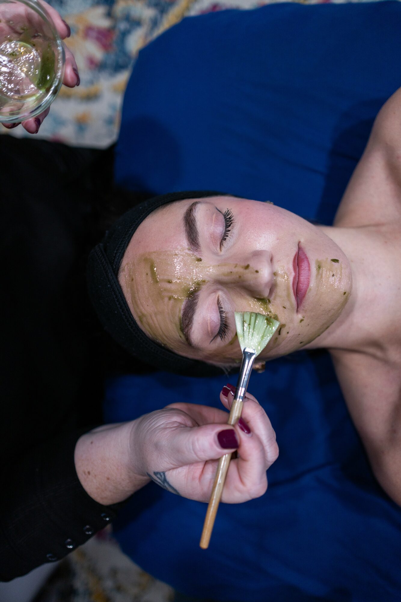 Woman with facial mask receiving a facial treatment, lying on a blue surface with eyes closed.