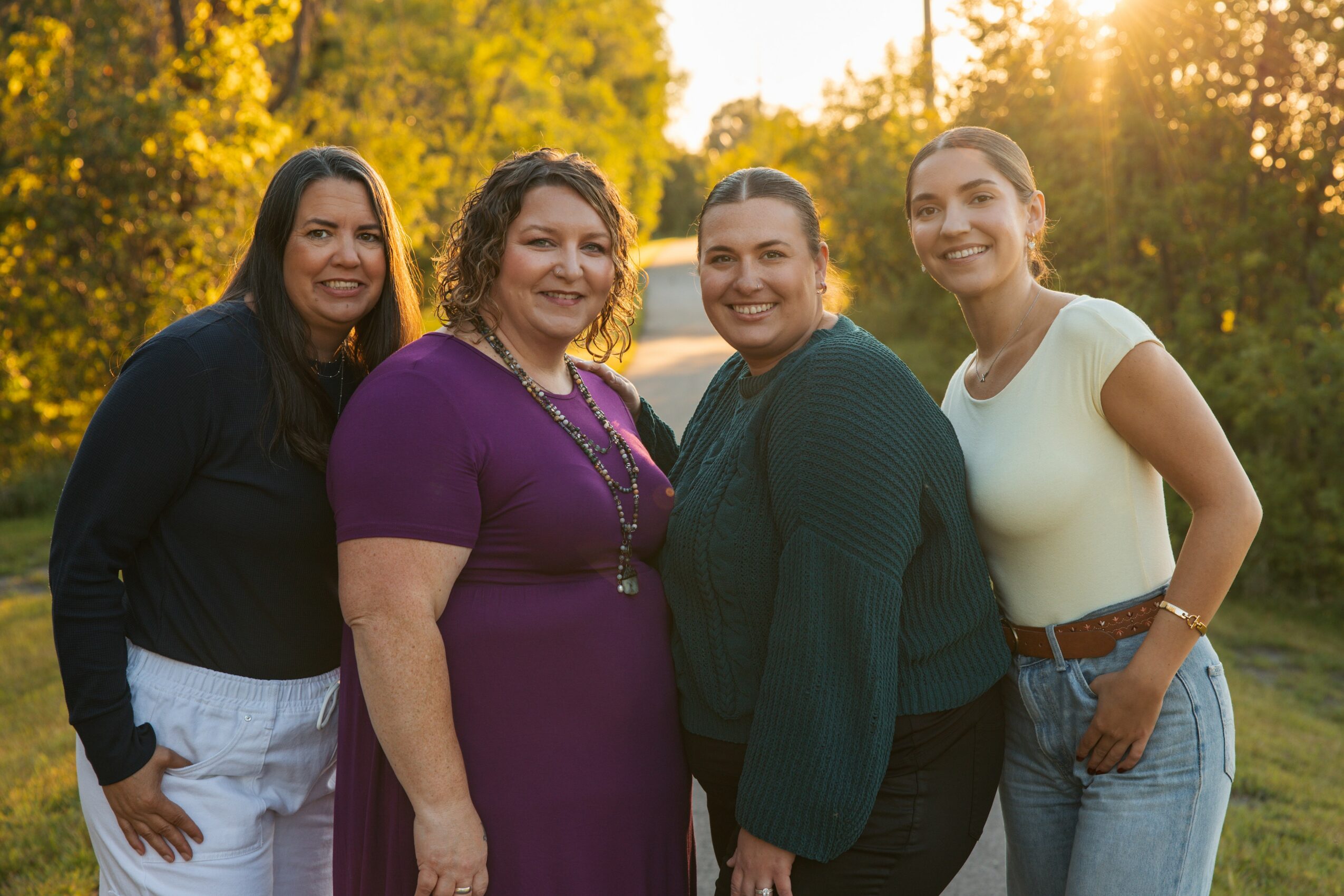 Four women standing outdoors in a park during sunset, smiling, with trees in the background.
