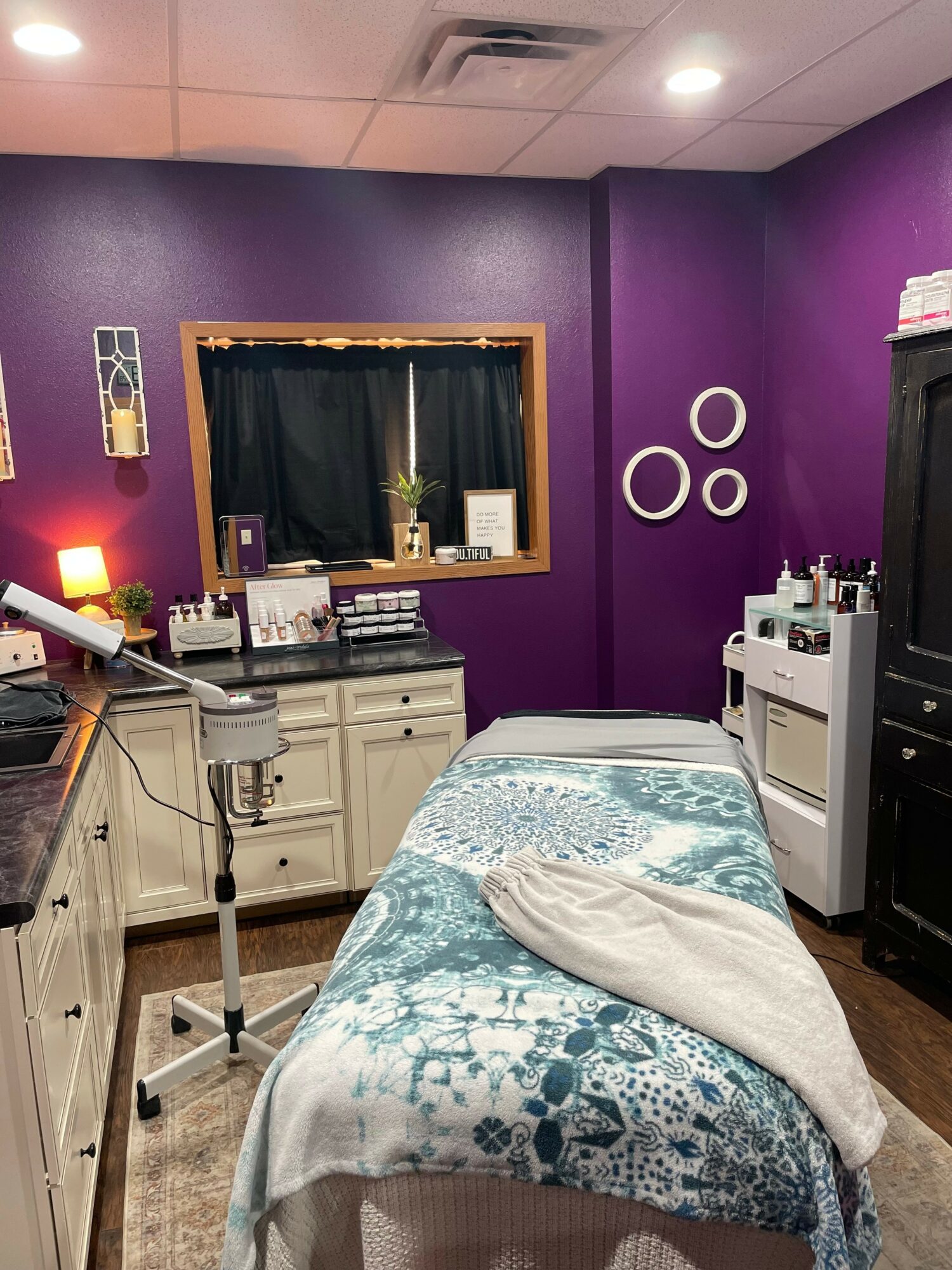 Treatment room with purple walls, bed with patterned blanket, window with black curtain, and medical equipment.