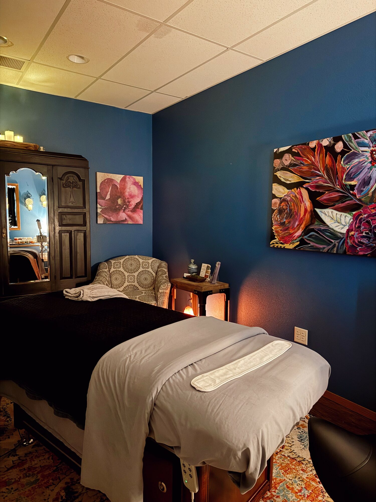 Bedroom with blue walls, bed with white and black bedding, colorful artwork, wooden wardrobe, and small side table.