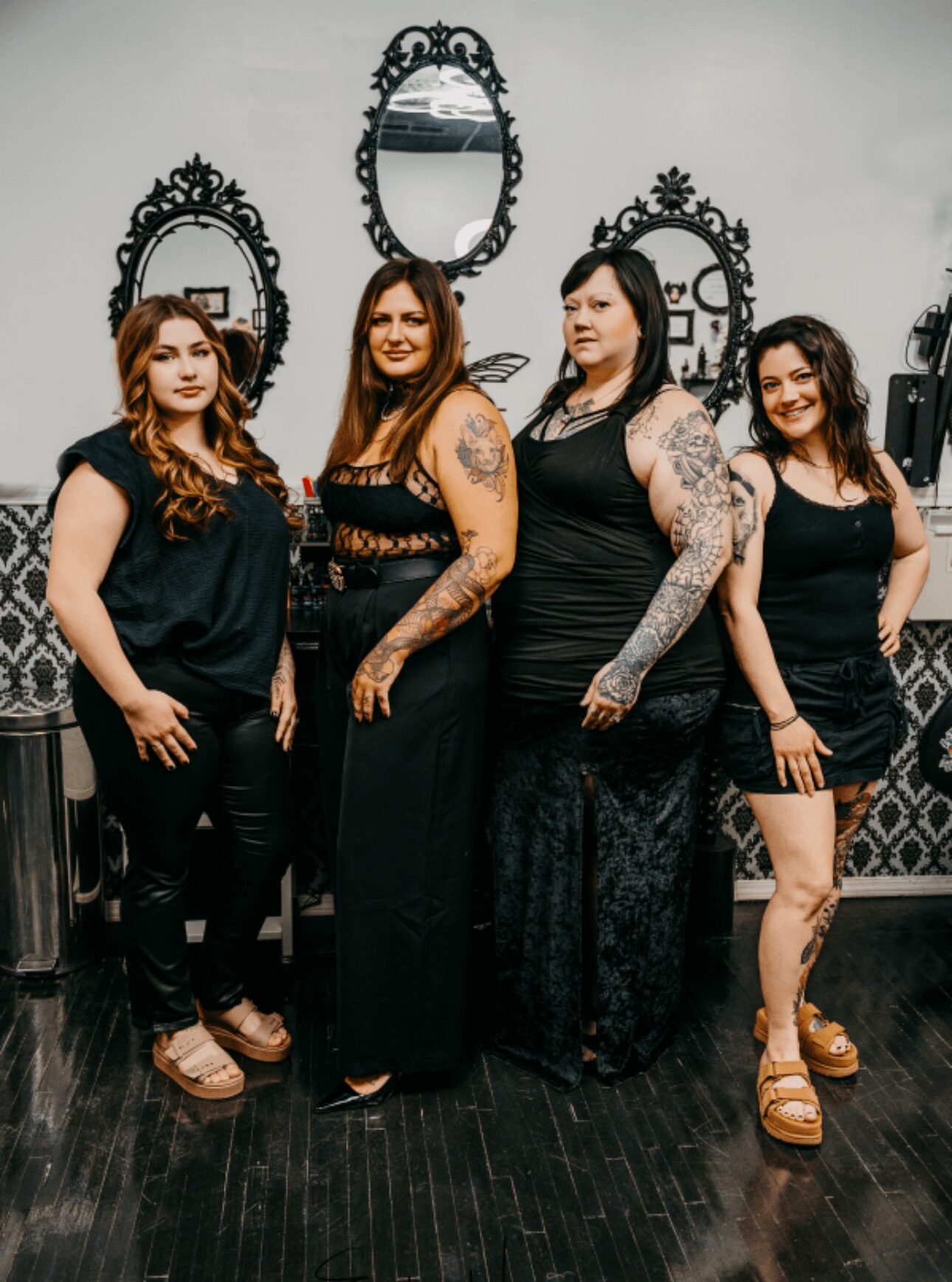 Four women standing together in front of ornate mirrors on a wall, dressed in black outfits, smiling.