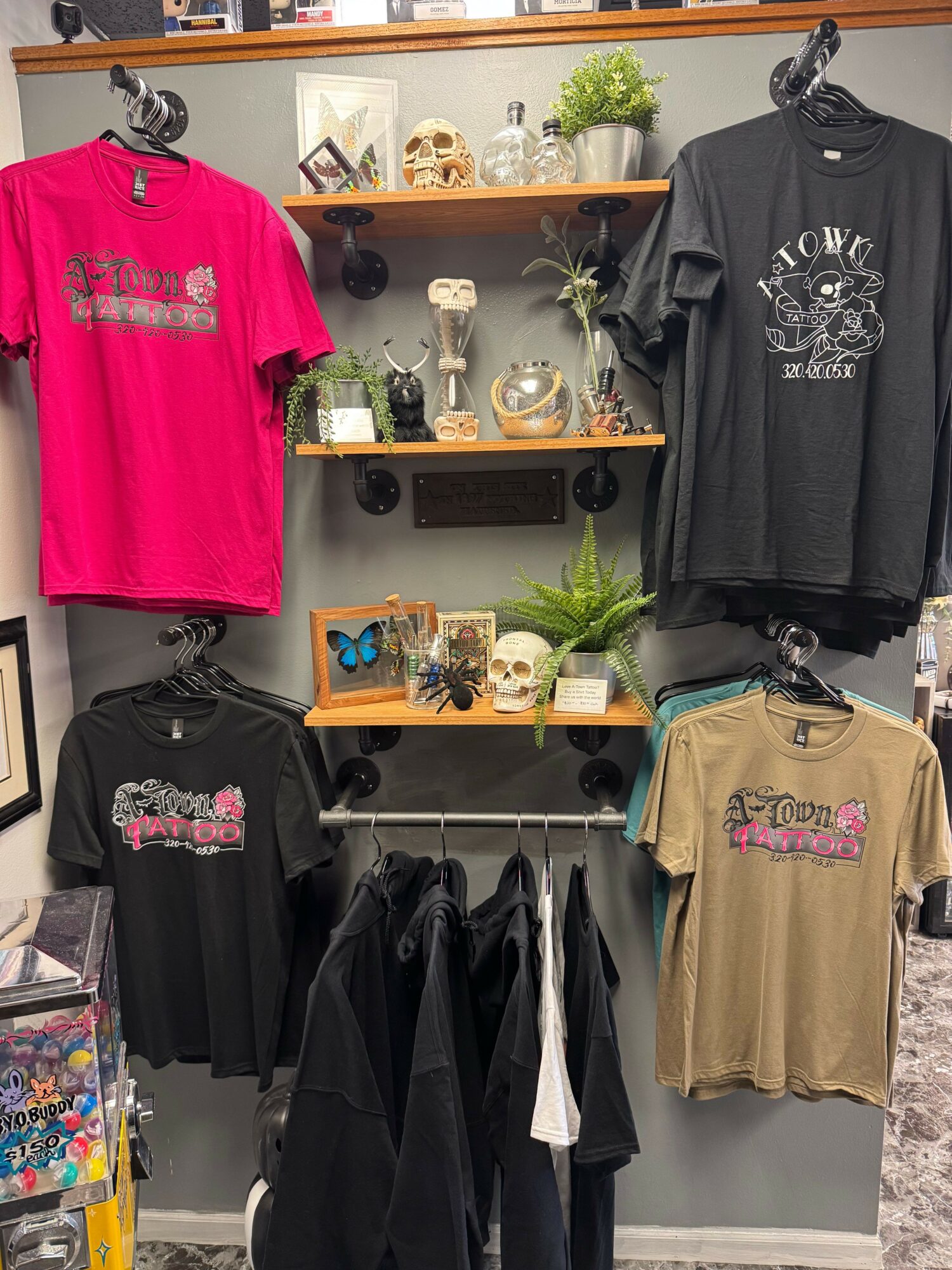 Display of four T-shirts on a wall with shelves and decorations behind, including plants, skulls, and framed pictures.