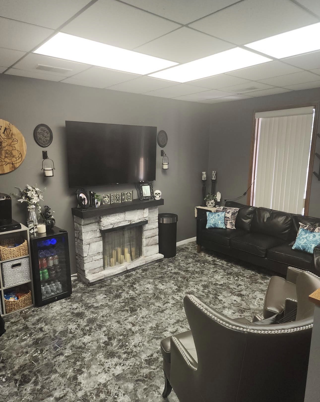 Living room with a large TV, fireplace, black sofa, armchair, and a window with blinds.
