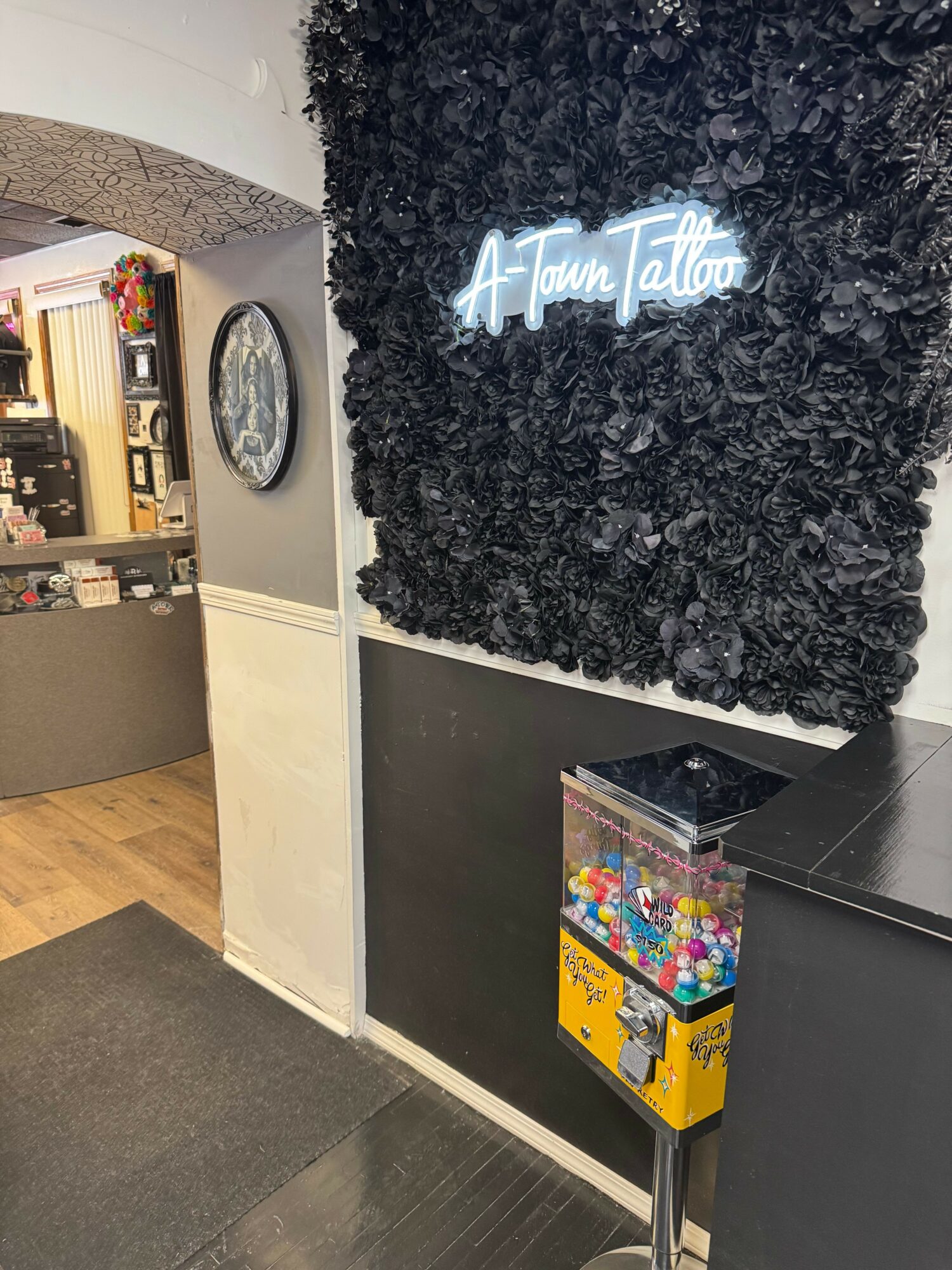 Neon sign reading 'A Town Tattoo' on a black floral wall, with a gumball machine below, in an indoor setting.