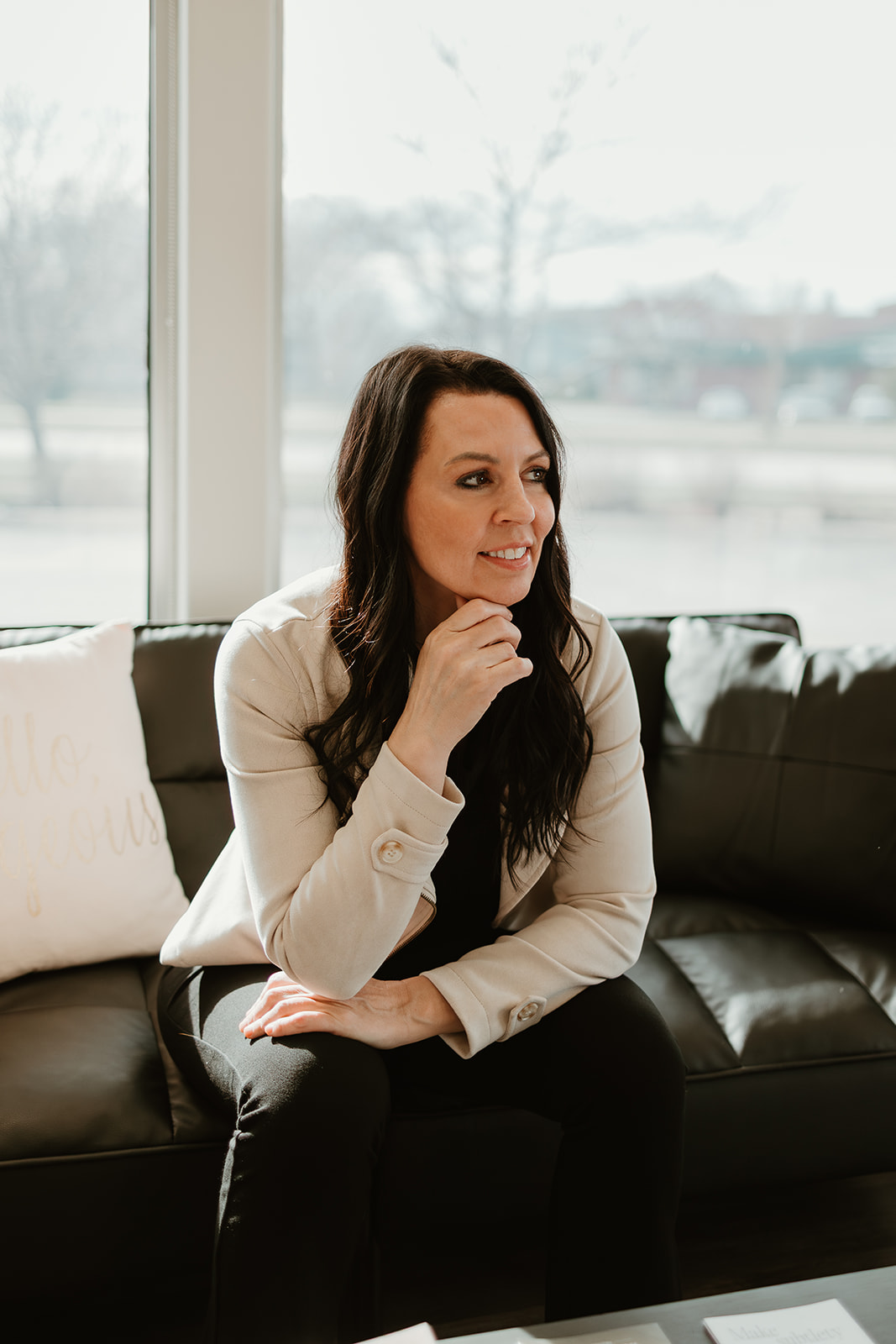 Woman with long dark hair sitting on a black sofa near a large window, looking to the side, smiling slightly.