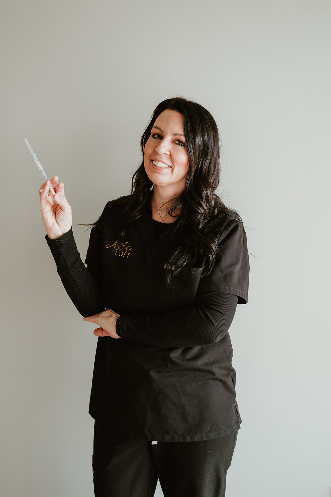 Woman with dark hair smiling, holding a dental tool, wearing black scrubs, standing against a plain light background.