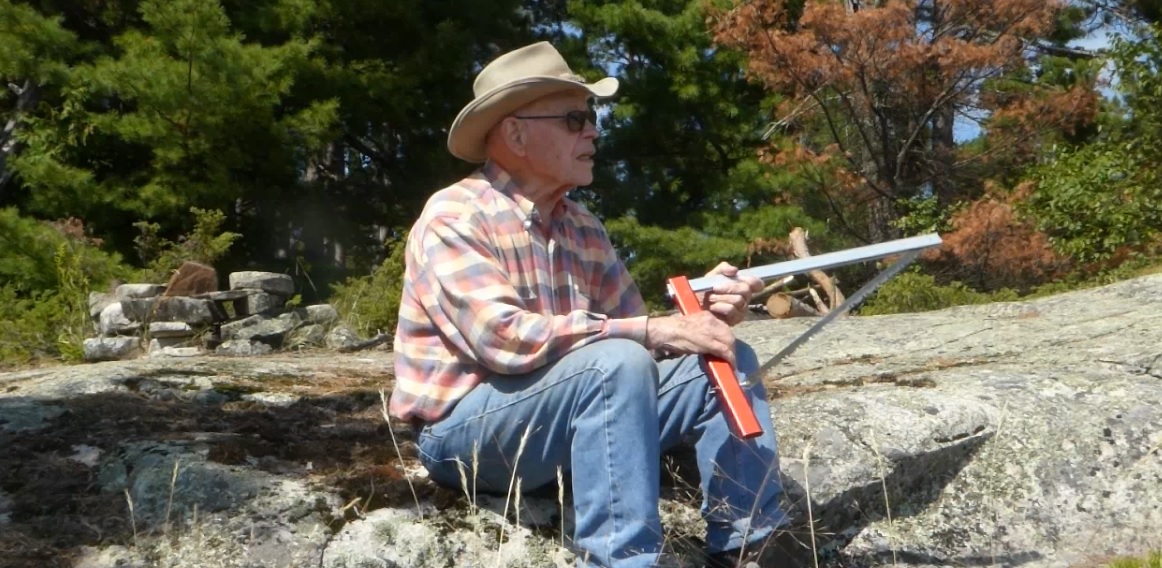 Elderly person sitting on rocks outdoors, fishing with a rod, wearing a hat and plaid shirt.