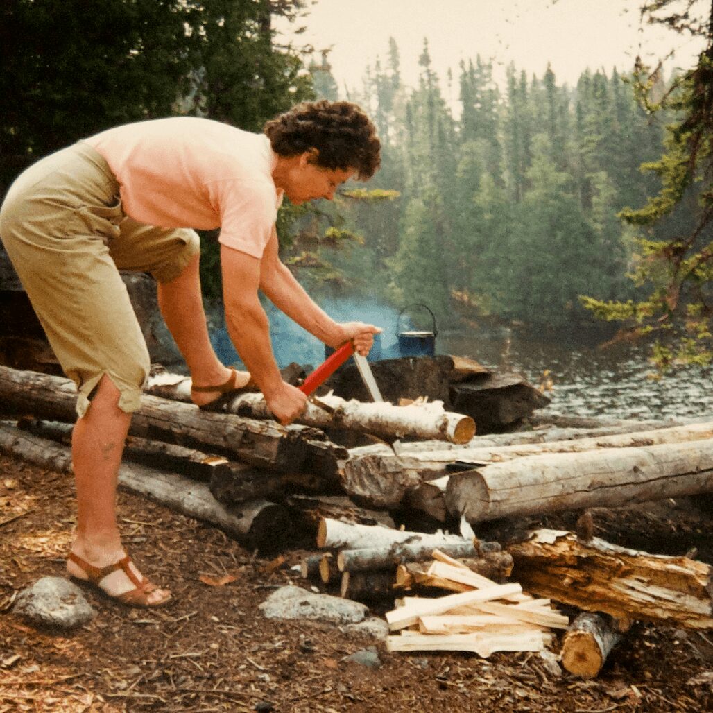 Person cutting wood with a saw near a lake surrounded by trees, wearing a pink shirt and beige shorts.