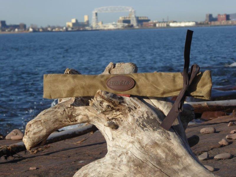 A piece of driftwood with a paper package and black ribbon on a rocky shoreline, water and cityscape in background.
