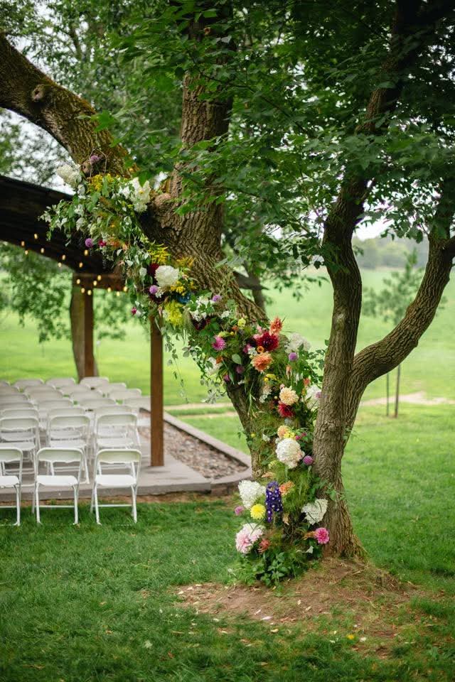 Tree decorated with colorful flowers and greenery, outdoor setting with chairs nearby, lush grass and trees in background.
