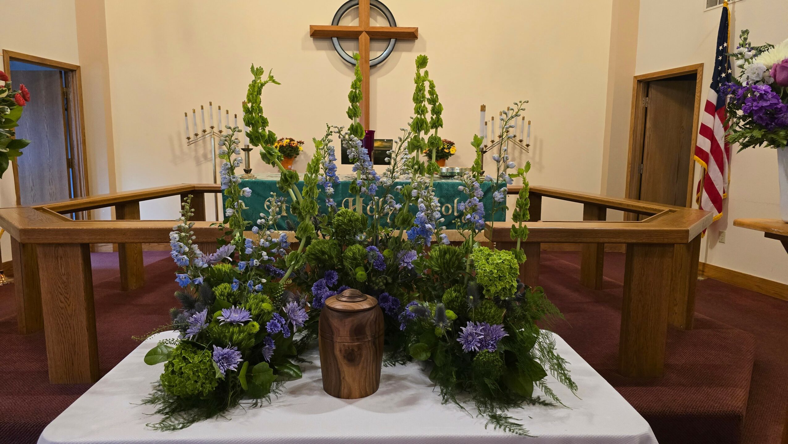 Floral arrangement with purple and green flowers on a table in front of a cross and chairs in a church setting.
