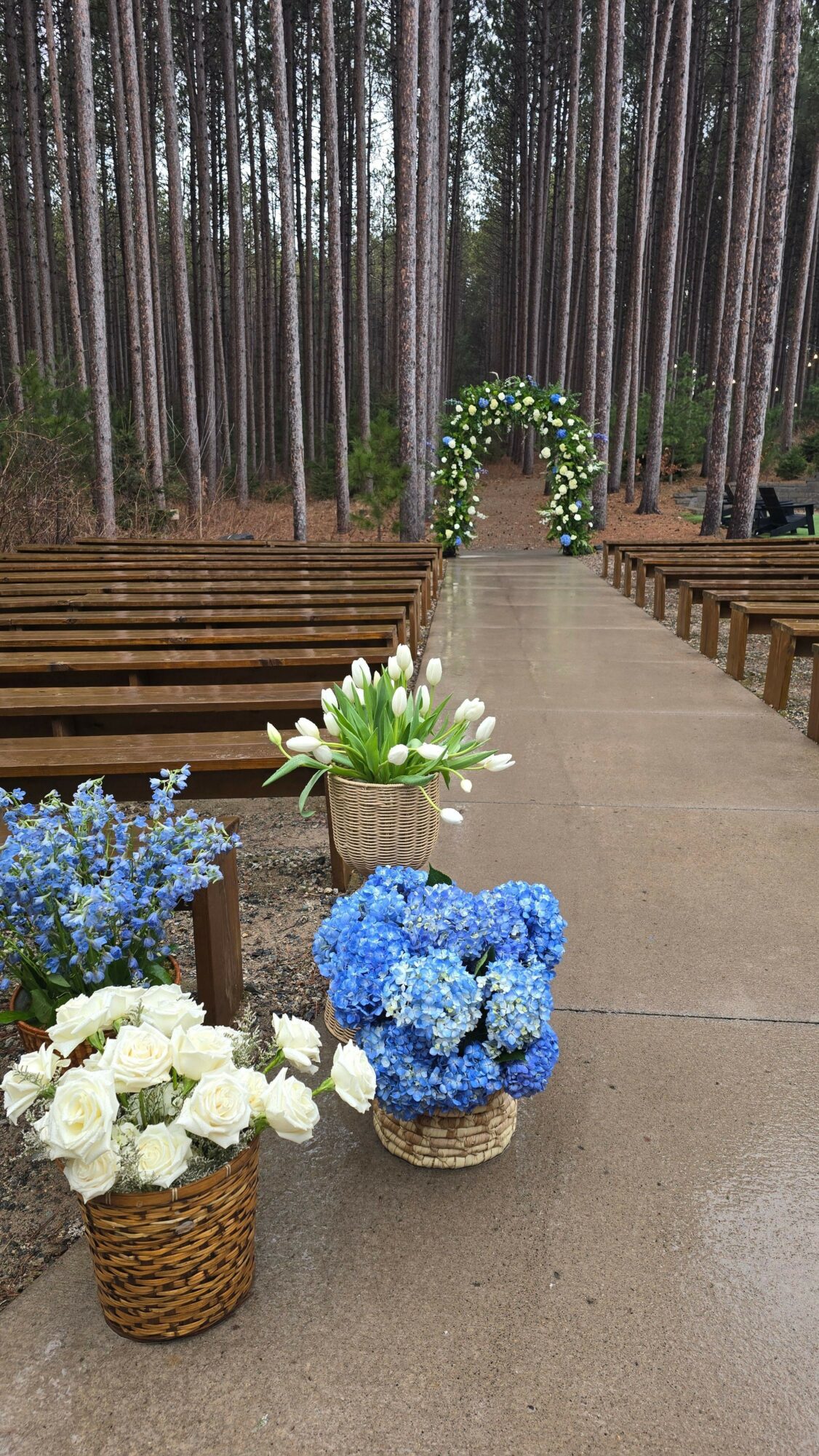 Outdoor wedding setup with flower arrangements and an arch in a forest setting, benches on both sides of a central aisle.