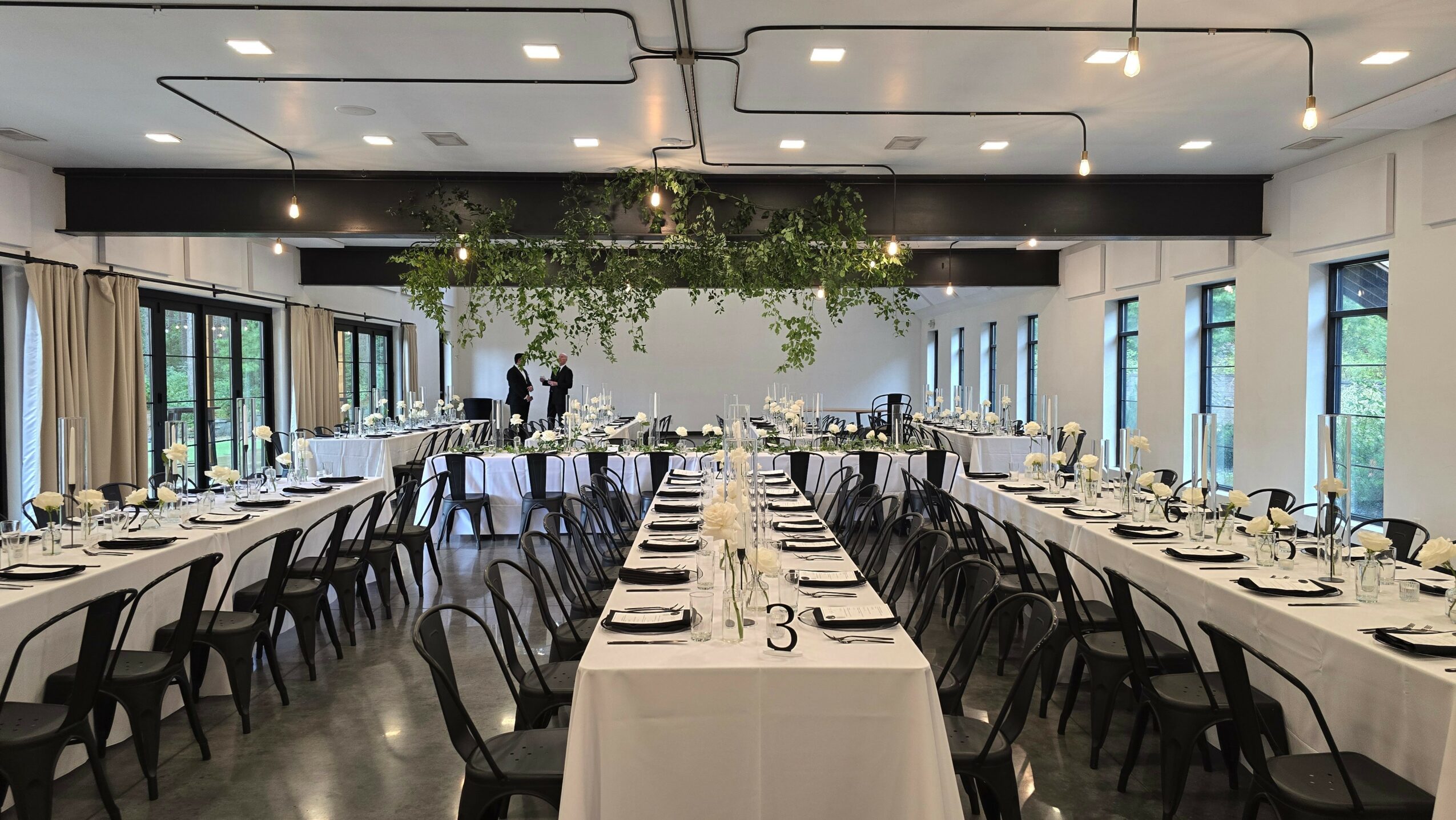Elegant banquet hall with long tables, chairs, table settings, and hanging greenery decoration, large windows on sides, modern ceiling design.