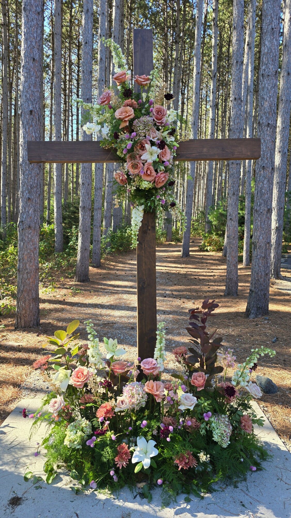 Wooden cross decorated with pink and white flowers, surrounded by a floral arrangement at the base, in a forest setting.