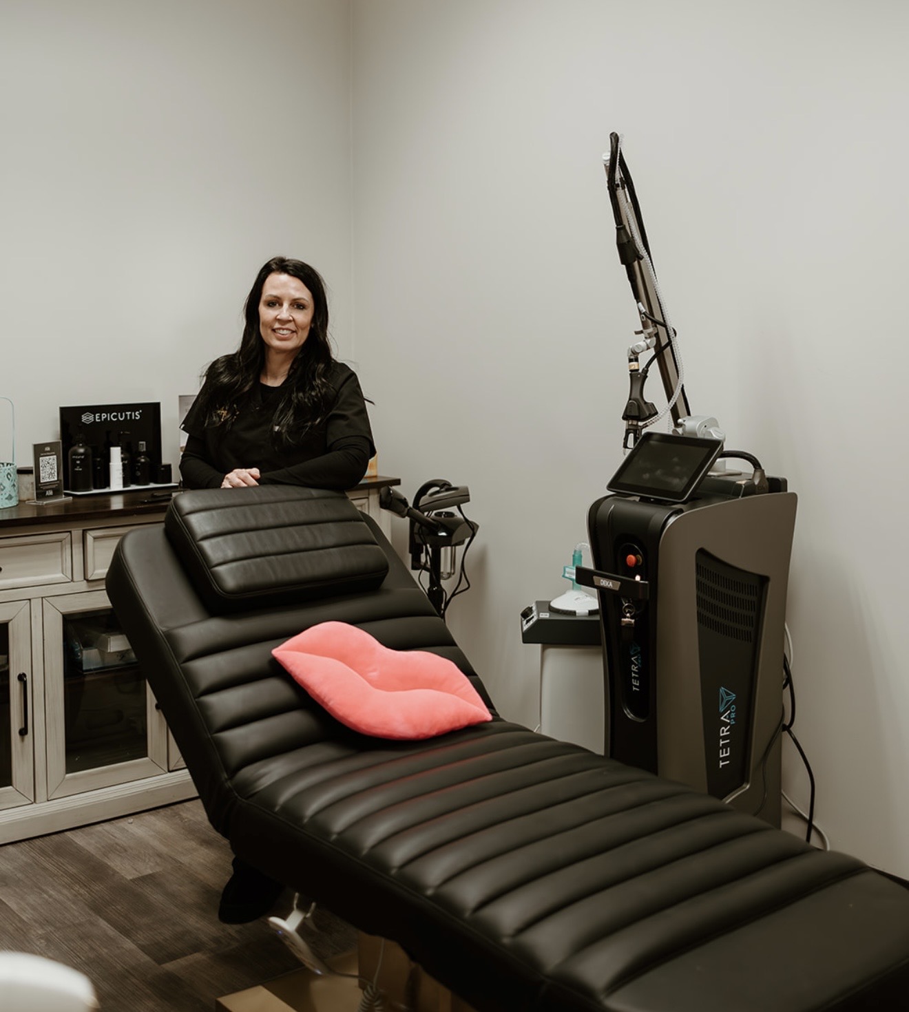 Woman standing in a medical or spa room with a treatment bed, medical equipment, and a pillow on the bed.