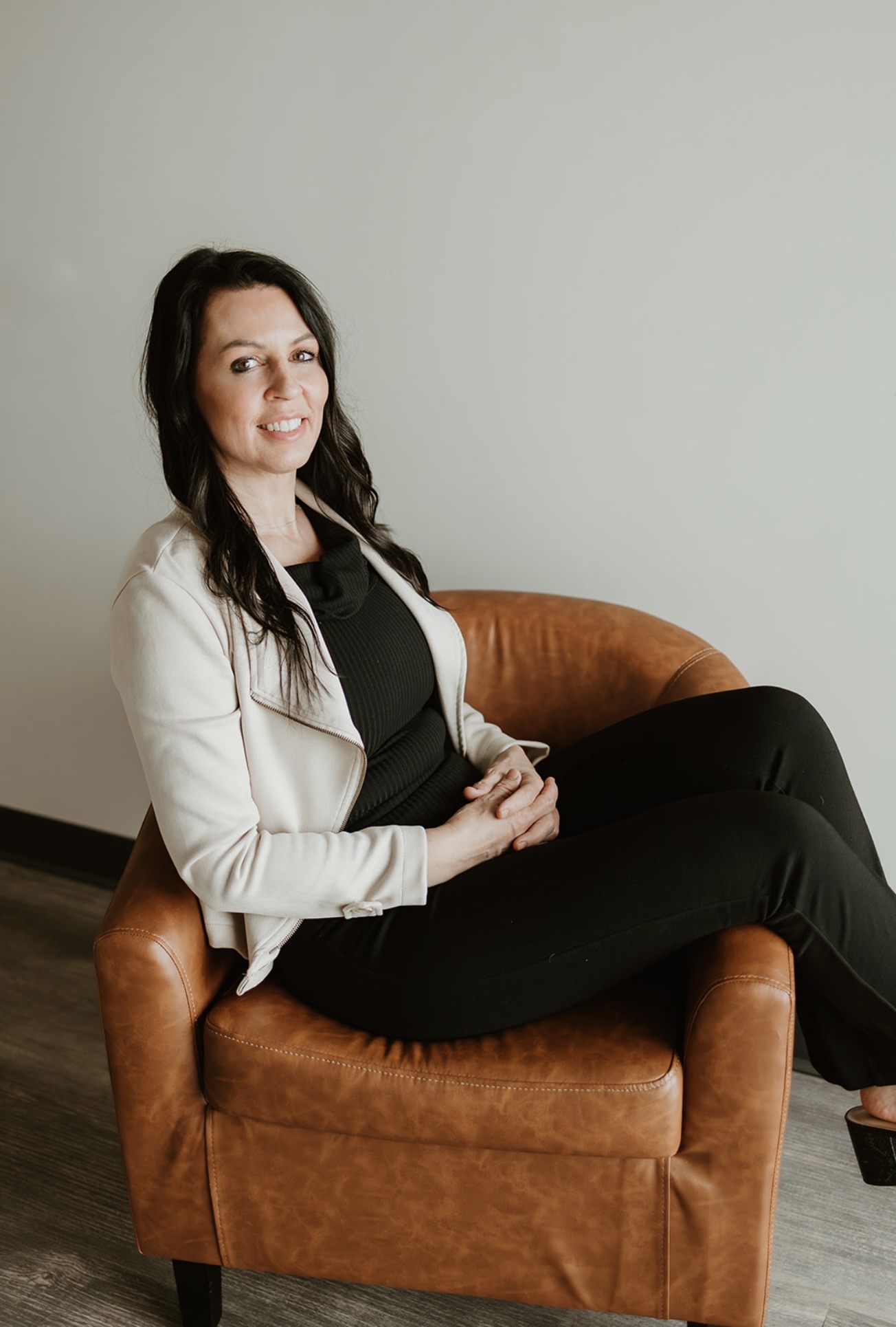 Woman sitting on a brown armchair, smiling, wearing a white blazer and black outfit, against a plain light-colored wall.