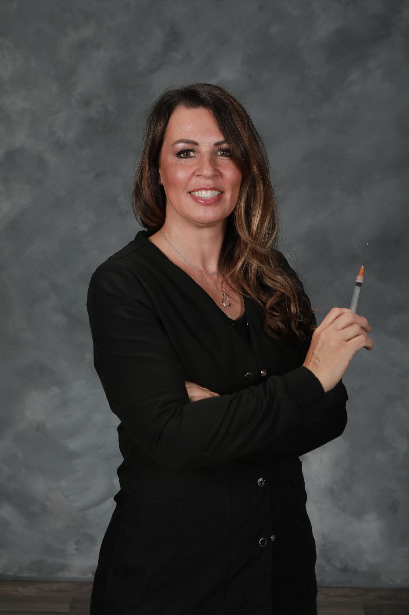 Woman with long wavy hair smiling, holding a pen, wearing a black top, standing against a gray background.