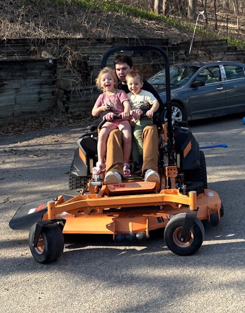 Three people sitting on a large orange lawn mower in a parking lot with a car and wooden fence in background.
