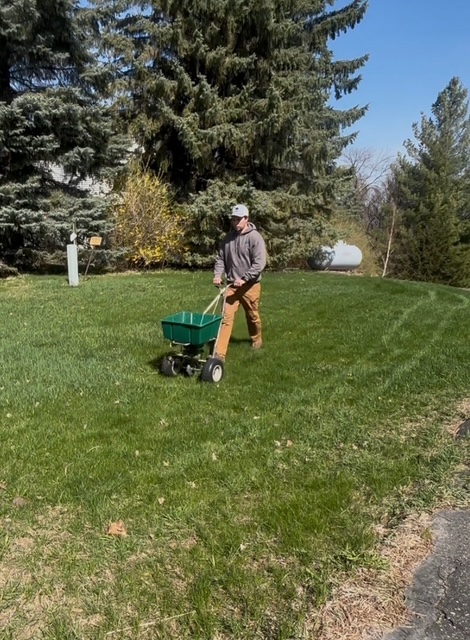 Person wearing a hat and gray shirt pushing a green wheelbarrow on grass with trees in background.