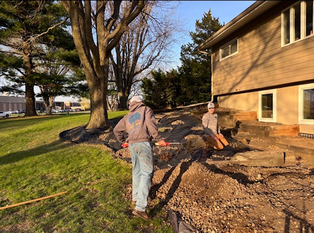 Two people working outdoors near a house, with trees and grass in the background.