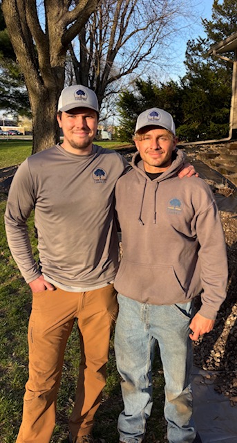 Two men standing outdoors, one with arm around the other, both wearing gray caps and casual clothing.
