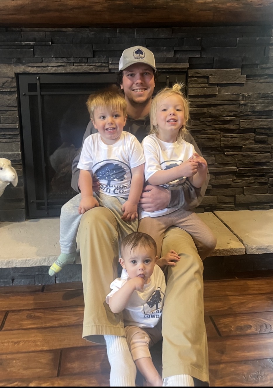 Person with three children sitting on a wooden floor in front of a stone fireplace, smiling.