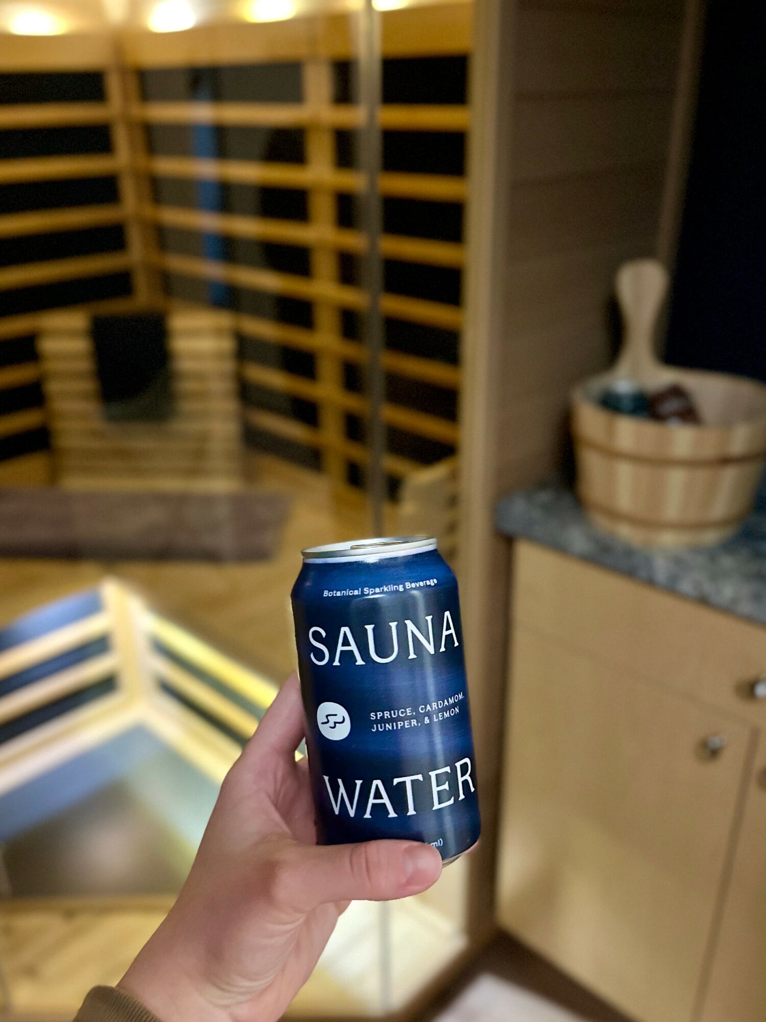 Hand holding a blue can labeled 'SAUNA WATER' in a kitchen with wooden cabinets and a countertop.