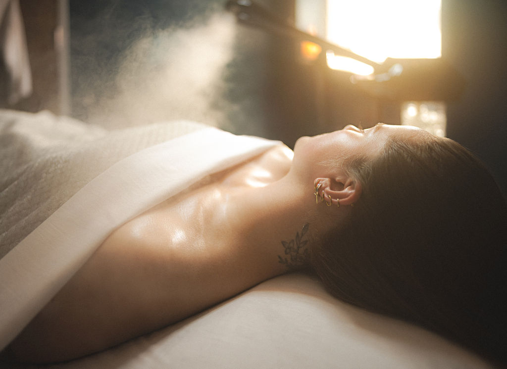 Person lying on bed with sunlight streaming through window, back and shoulder visible, relaxed posture.