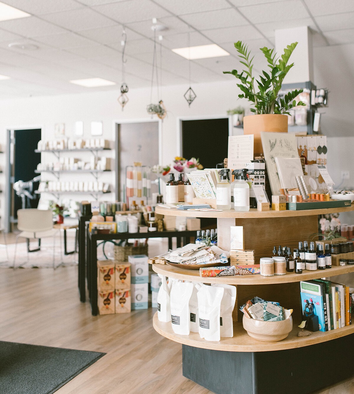 Interior of a store with shelves, a display table, and potted plants, featuring various products and a wooden floor.