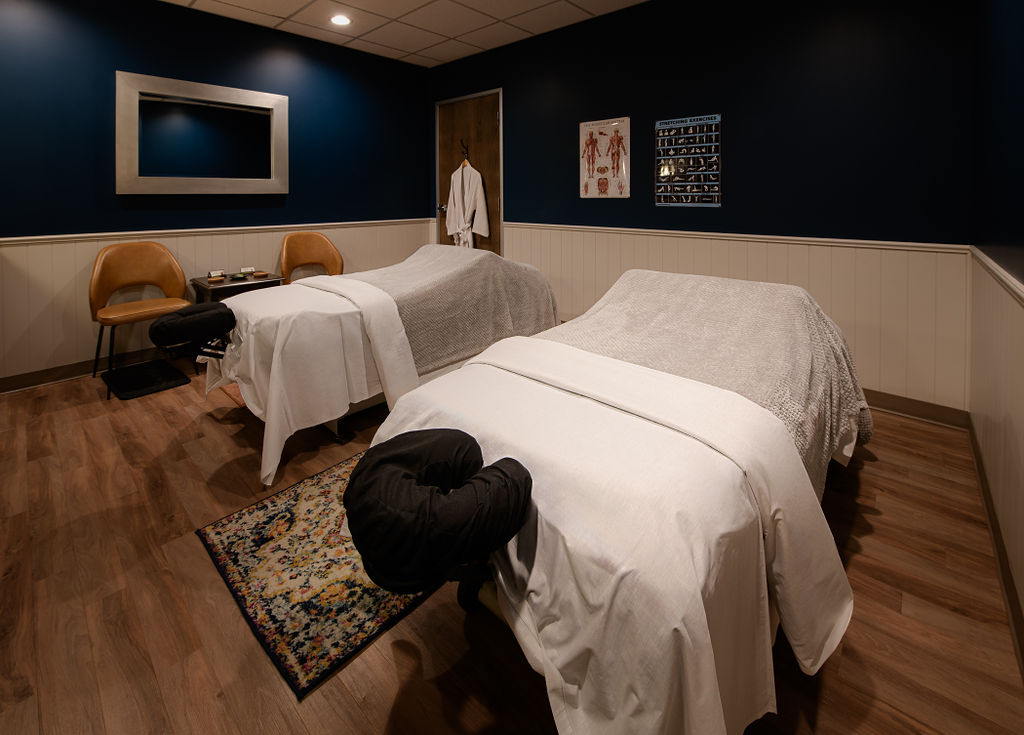 Two massage tables in a room with dark blue walls, framed pictures, and a wooden floor, with a rug and a coat hanger.